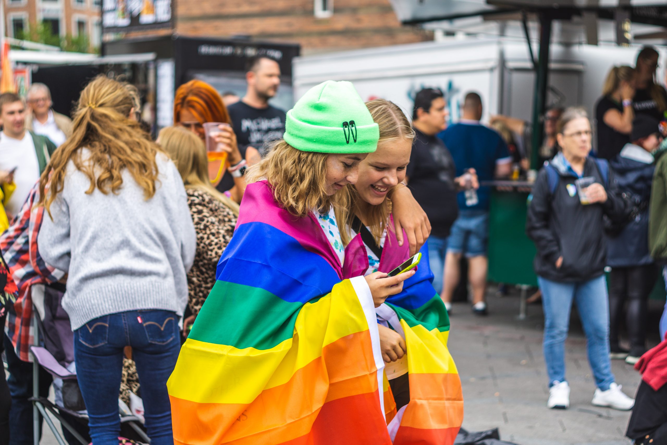 Two young female-presenting people walk down the street during pride festivals wrapped in a rainbow pride flag.