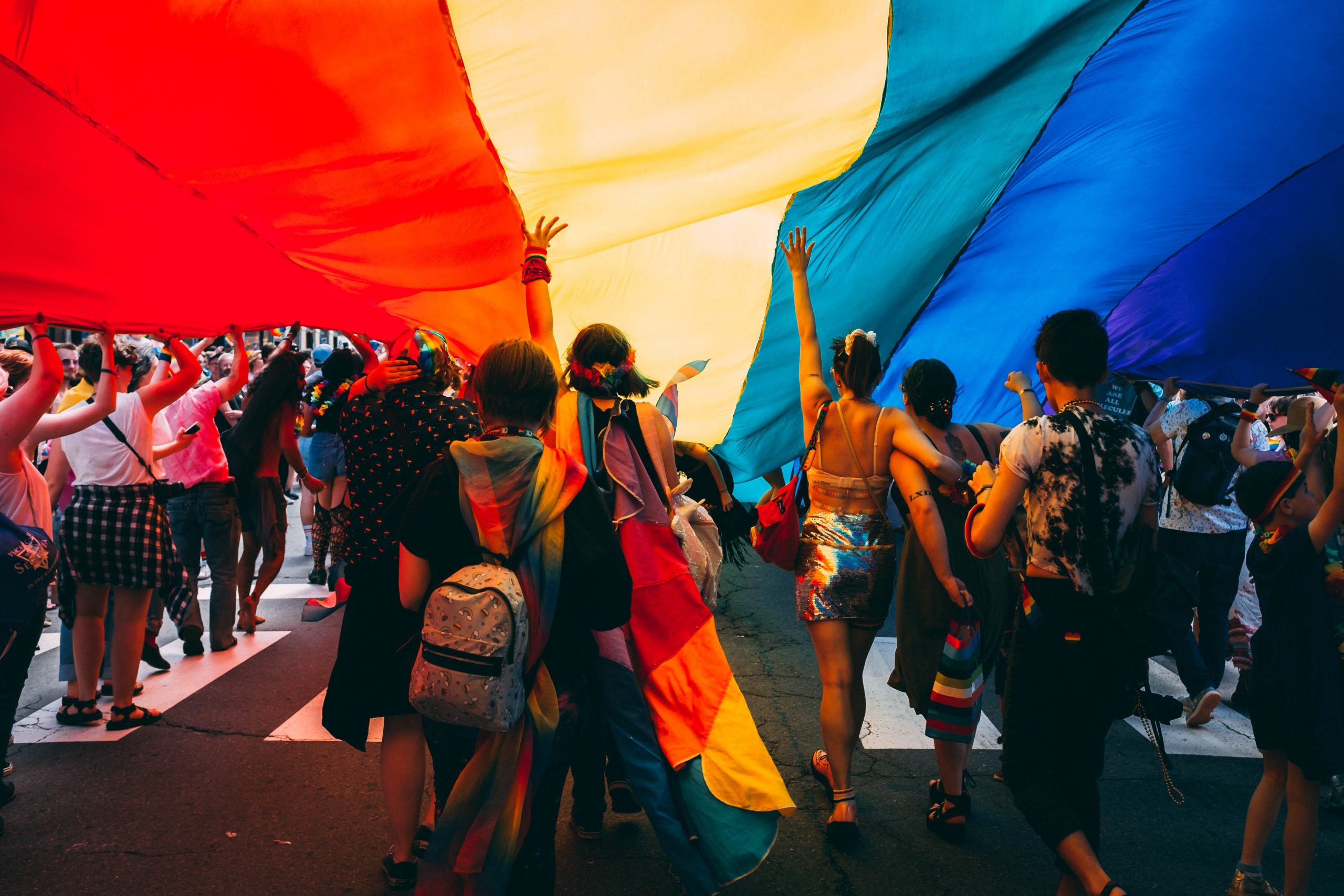 A group of individuals walk along the street underneath a giant rainbow pride flag at a pride festivals.