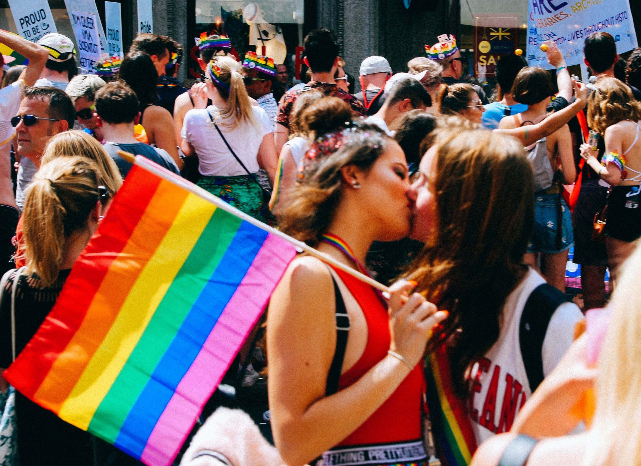 Two female-presenting people kiss in a busy crowd at a pride festival pride, while holding a rainbow pride flag.