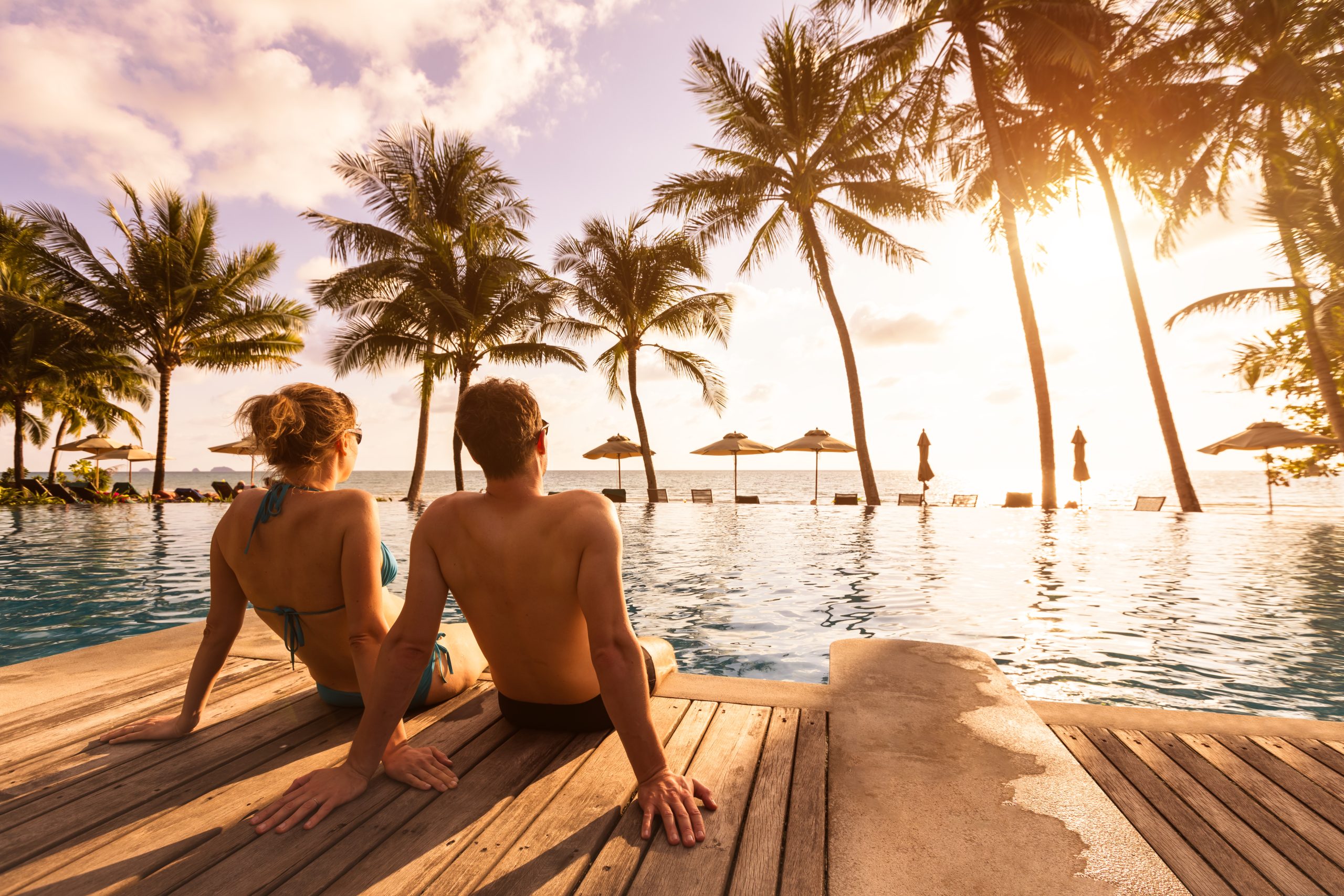 Couple sitting by resort pool at sunset, overlooking palm trees and the ocean.