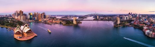 Pink sunrise over Sydney harbour, the harbour bridge and city CBDs on both shores in wide aerial panorama over calm waters disturbed by ferries and boats.
