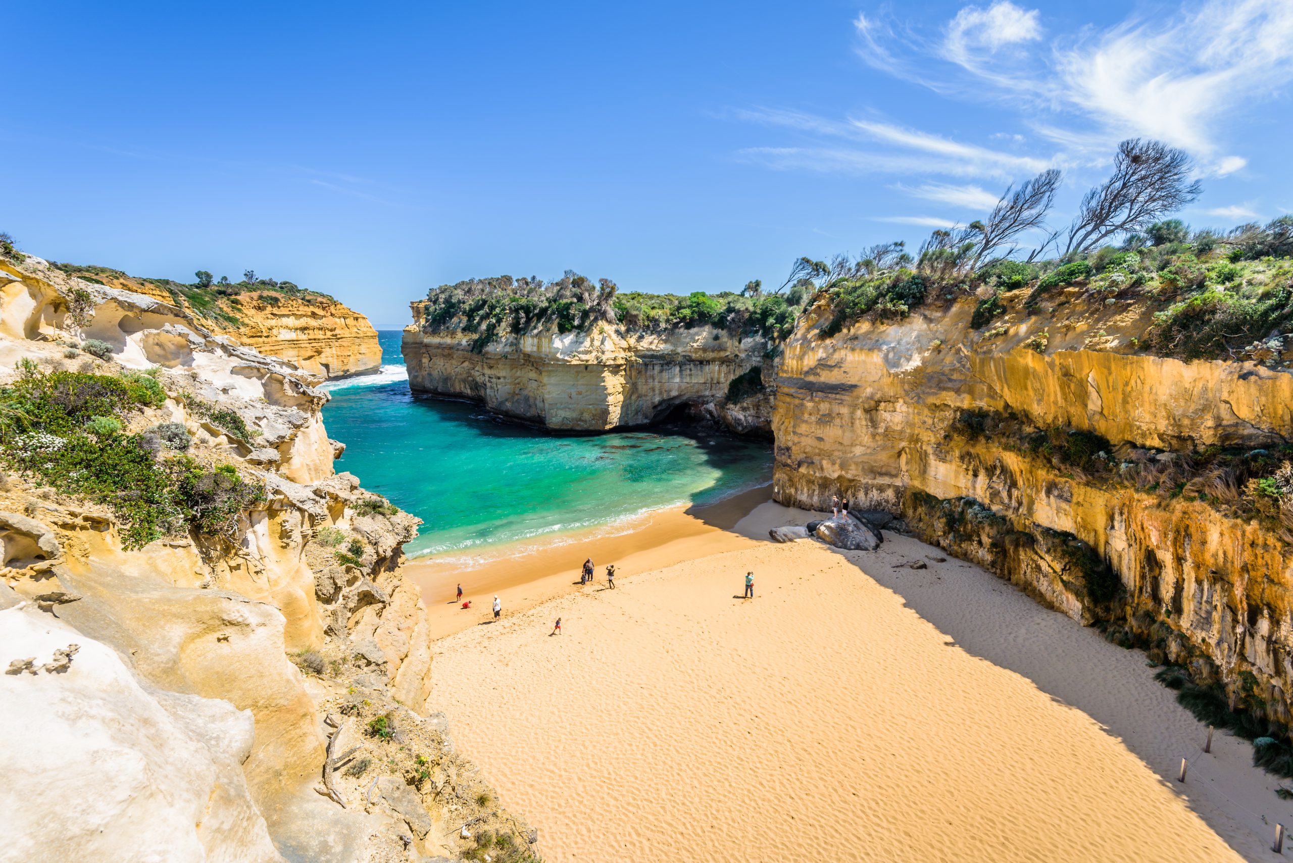 Loch Ard Gorge in Port Campbell National Park in Victoria, Australia on a bright sunny day. A few beachgoers and surfers sand on the sand on the waters edge.